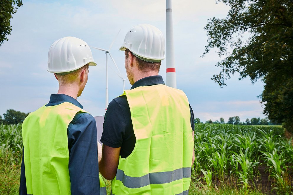 Zwei Personen mit Bauhelm und Warnweste stehen vor einem Windpark, man sieht die Personen von hinten