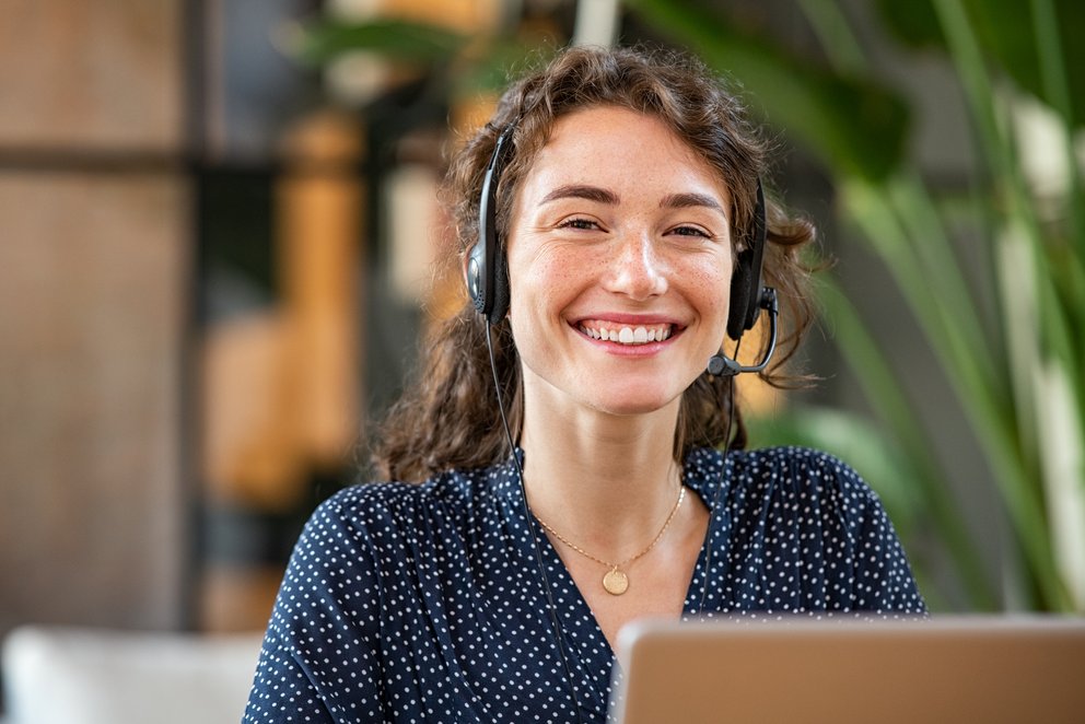 Eine lächelnde Frau mit Headset sitzt am Laptop.