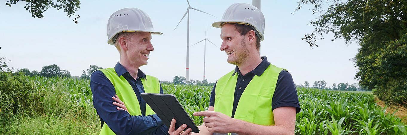 Zwei Personen mit Bauhelm und Warnweste stehen vor einem Windpark