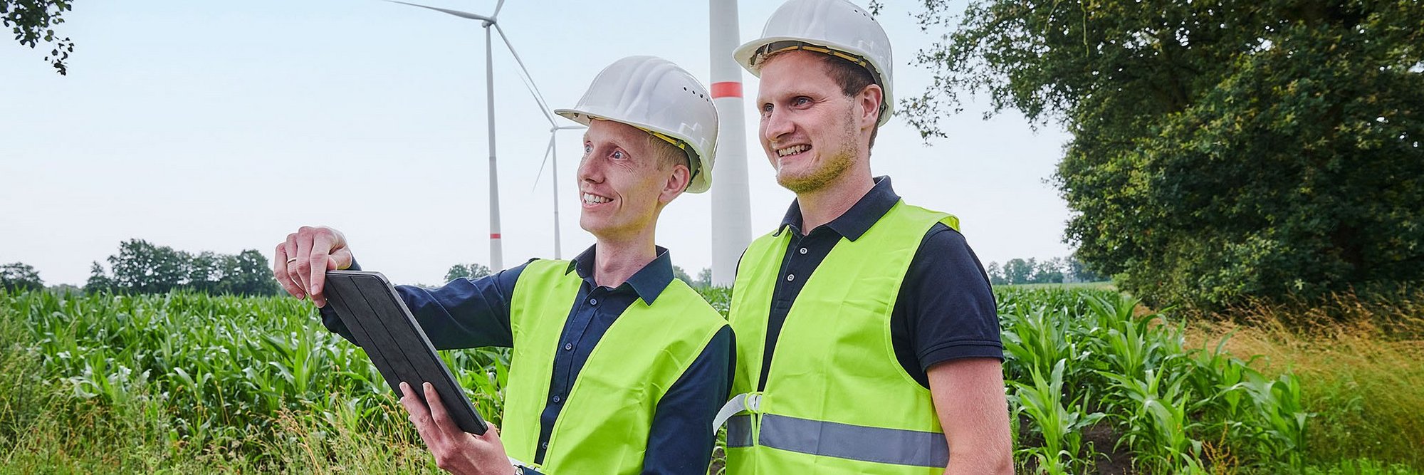Zwei Personen mit Bauhelm und Warnweste stehen vor einem Windpark