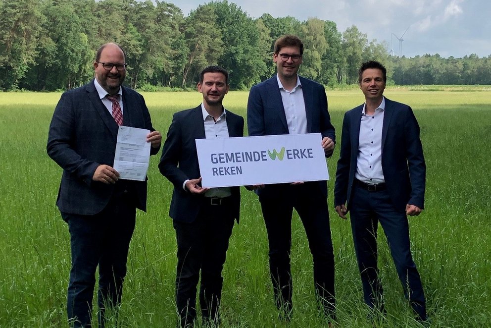 Gruppe von vier Männer in Anzügen mit Firmenschild im Grünen.
