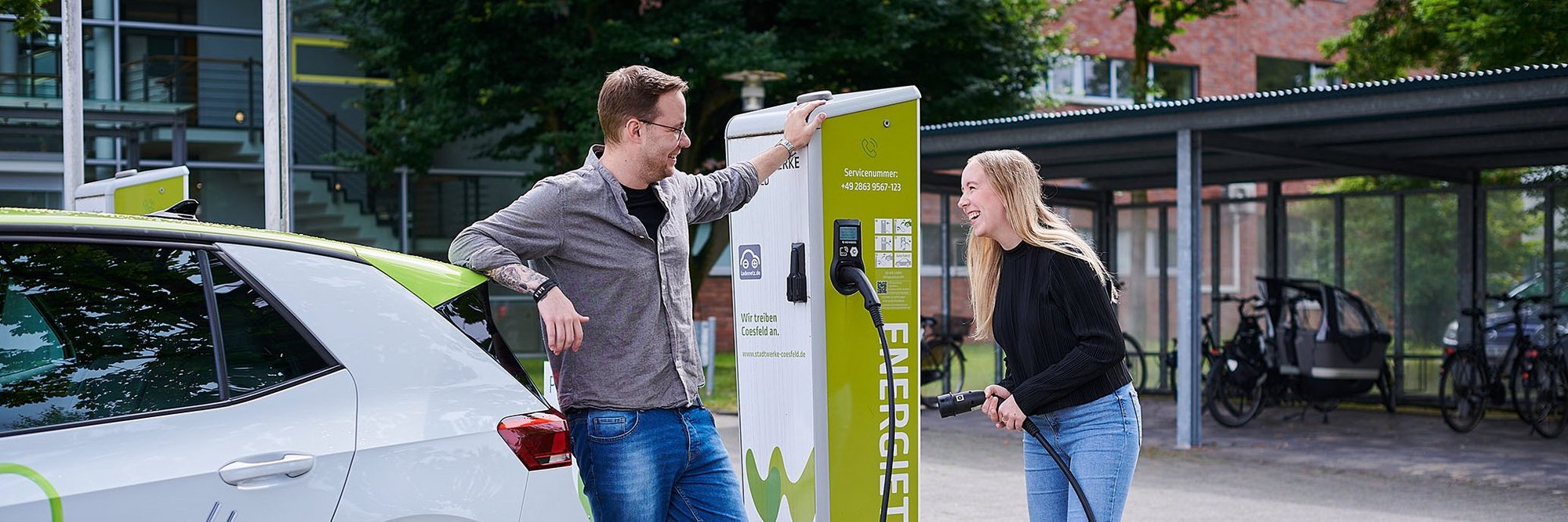 Eine Frau und ein Mann stehen mit einem Auto vor einer E-Ladesäule und tanken das Fahrzeug