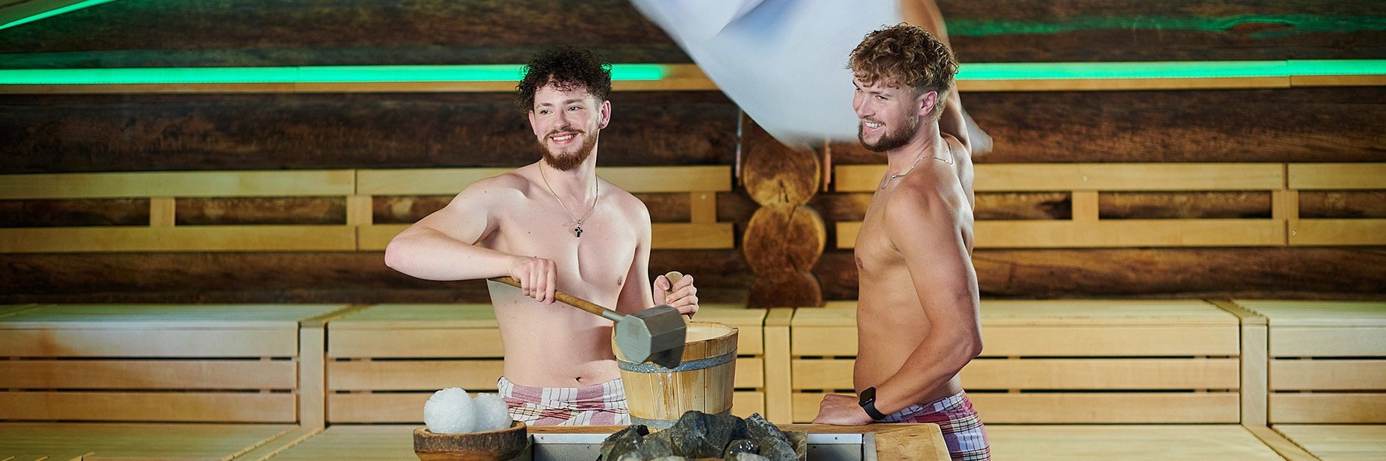 Zwei Männer mit freiem Oberkörper stehen in der Sauna hinter einem Saunaofen. Saunaaufguss