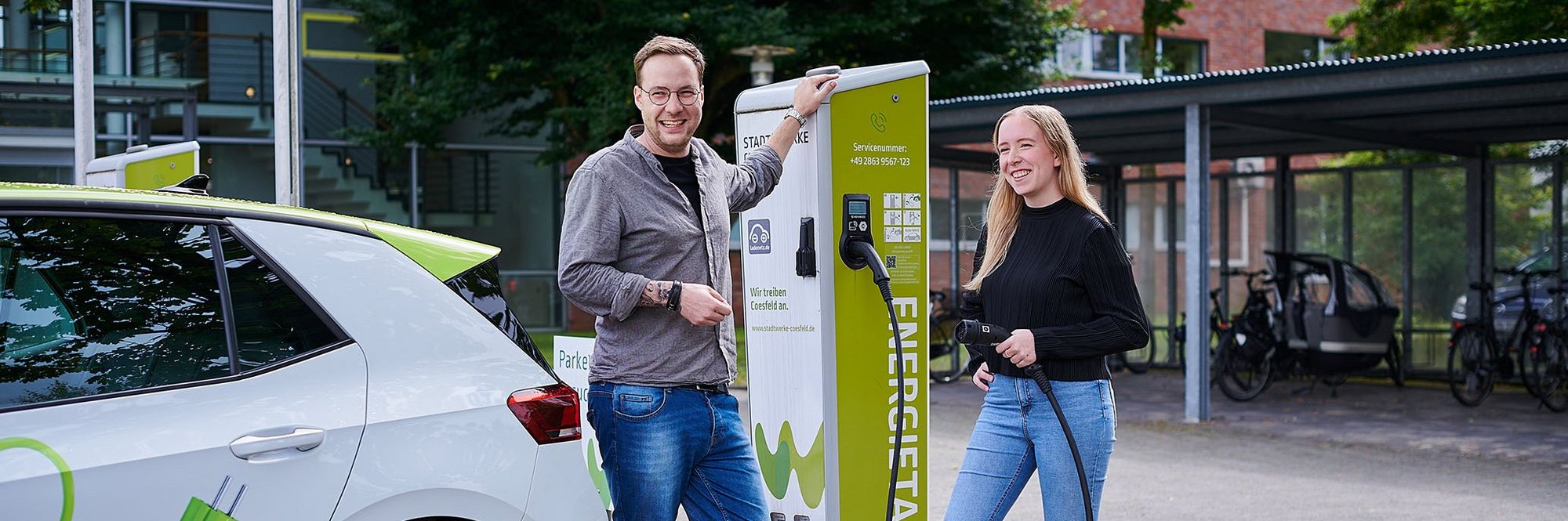 Eine Frau und ein Mann stehen mit einem Auto vor einer E-Ladesäule und tanken das Fahrzeug