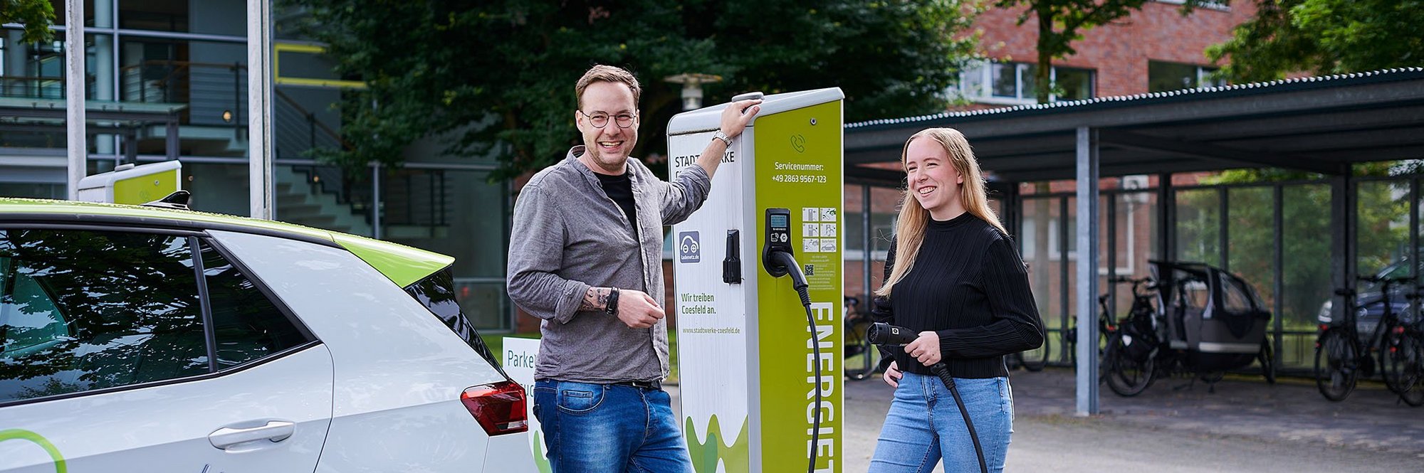 Eine Frau und ein Mann stehen mit einem Auto vor einer E-Ladesäule und tanken das Fahrzeug