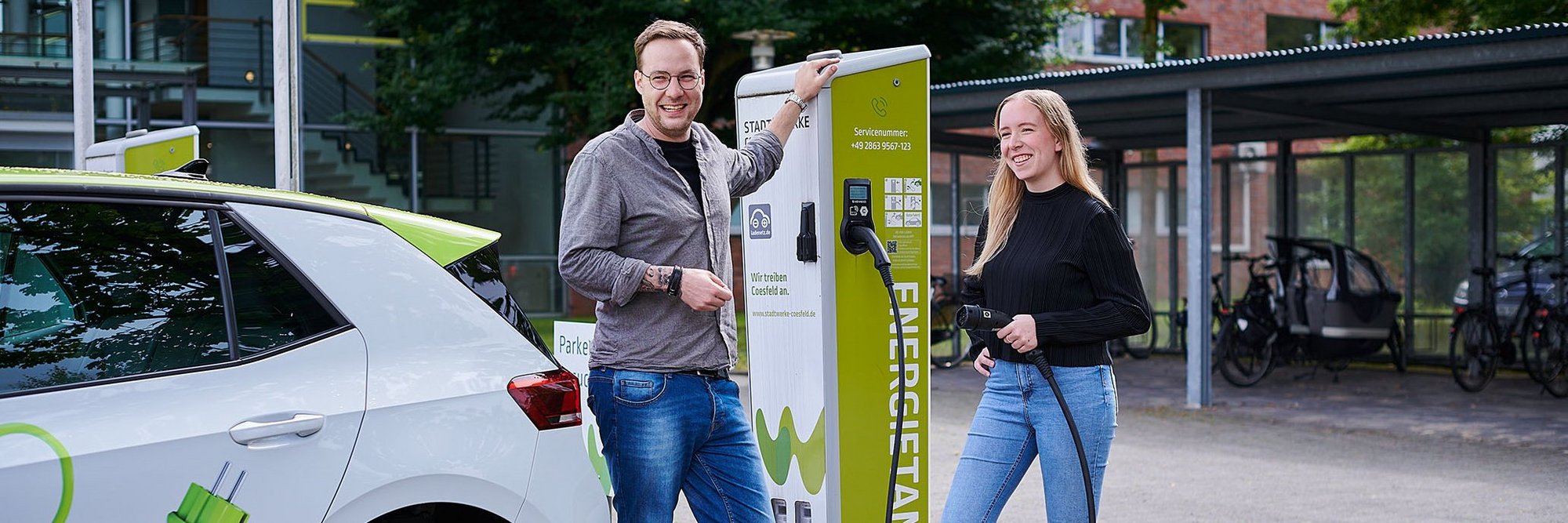 Eine Frau und ein Mann stehen mit einem Auto vor einer E-Ladesäule und tanken das Fahrzeug