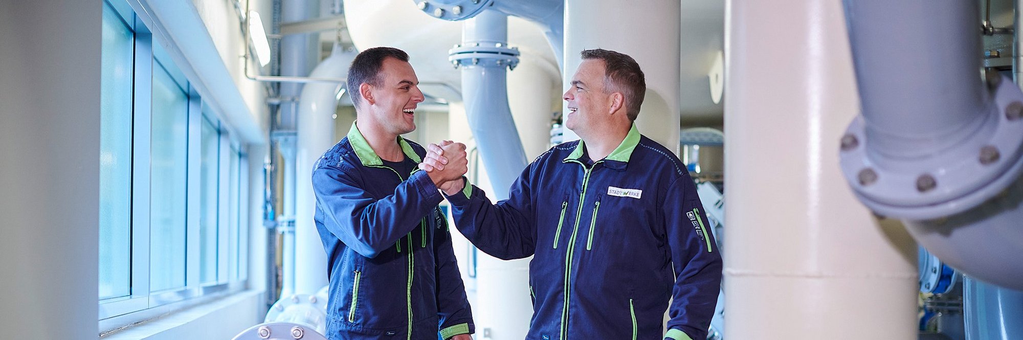 Zwei Techniker in Blaumann zwischen Wasserrohren, Handshake