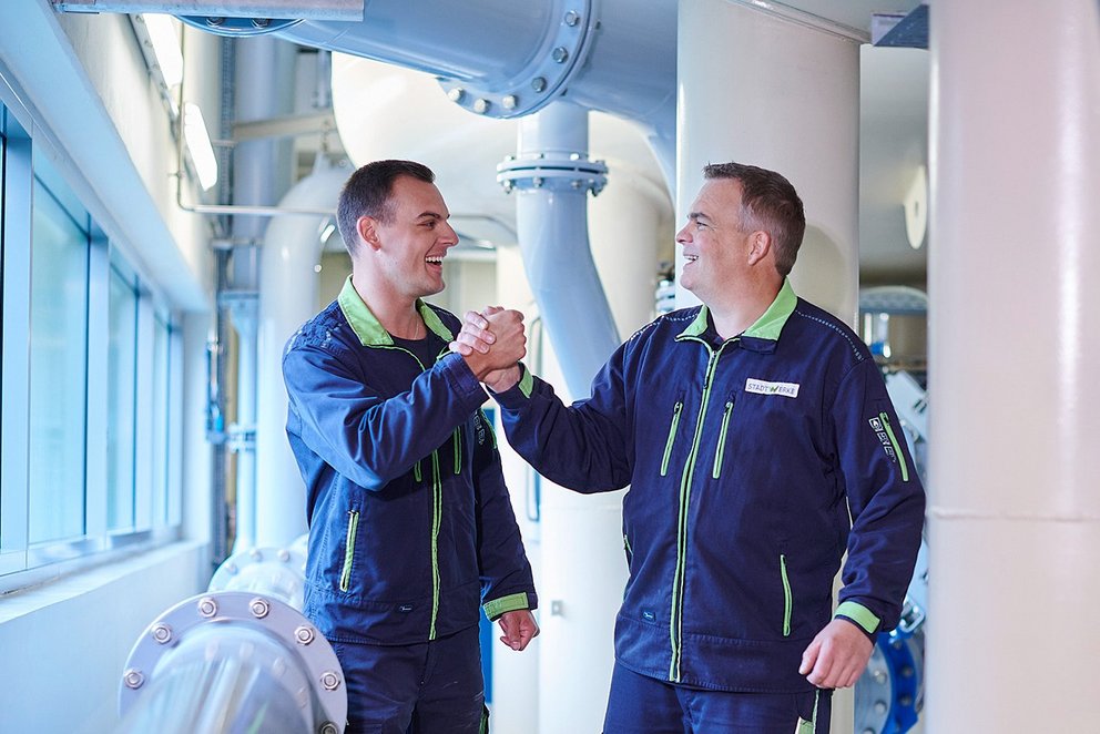 Zwei Techniker in Blaumann zwischen Wasserrohren, Handshake